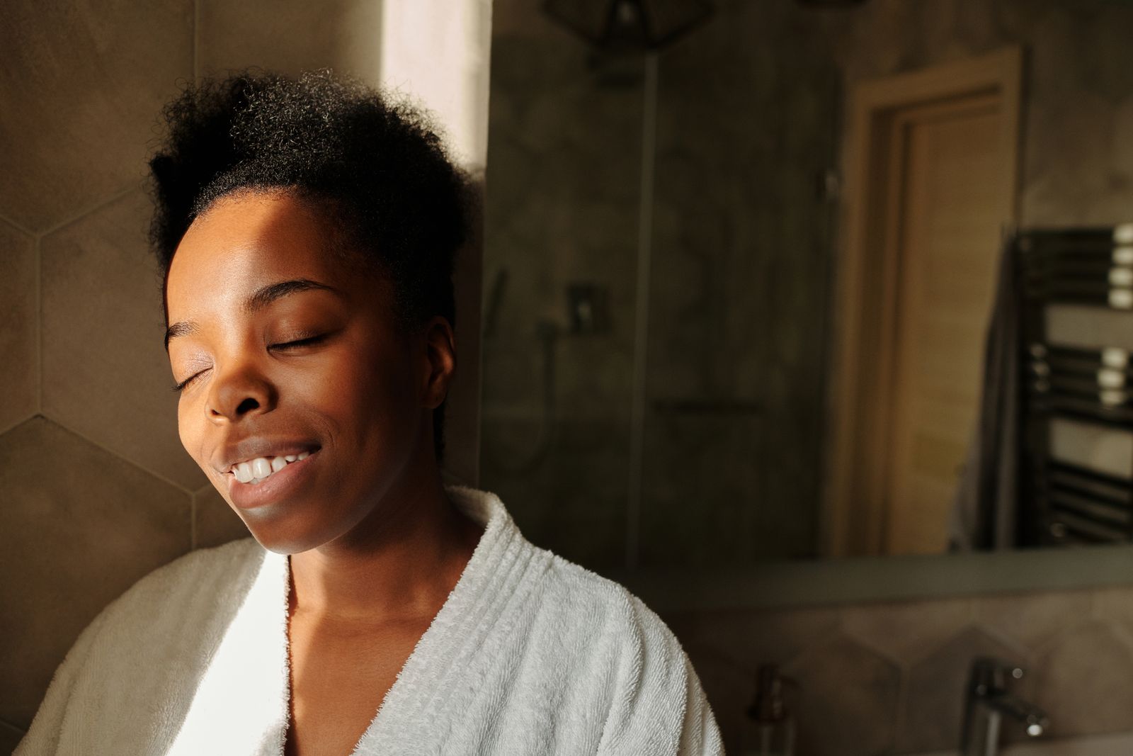 A woman in a bathrobe with her eyes closed, in a warm bathroom at the end of the day